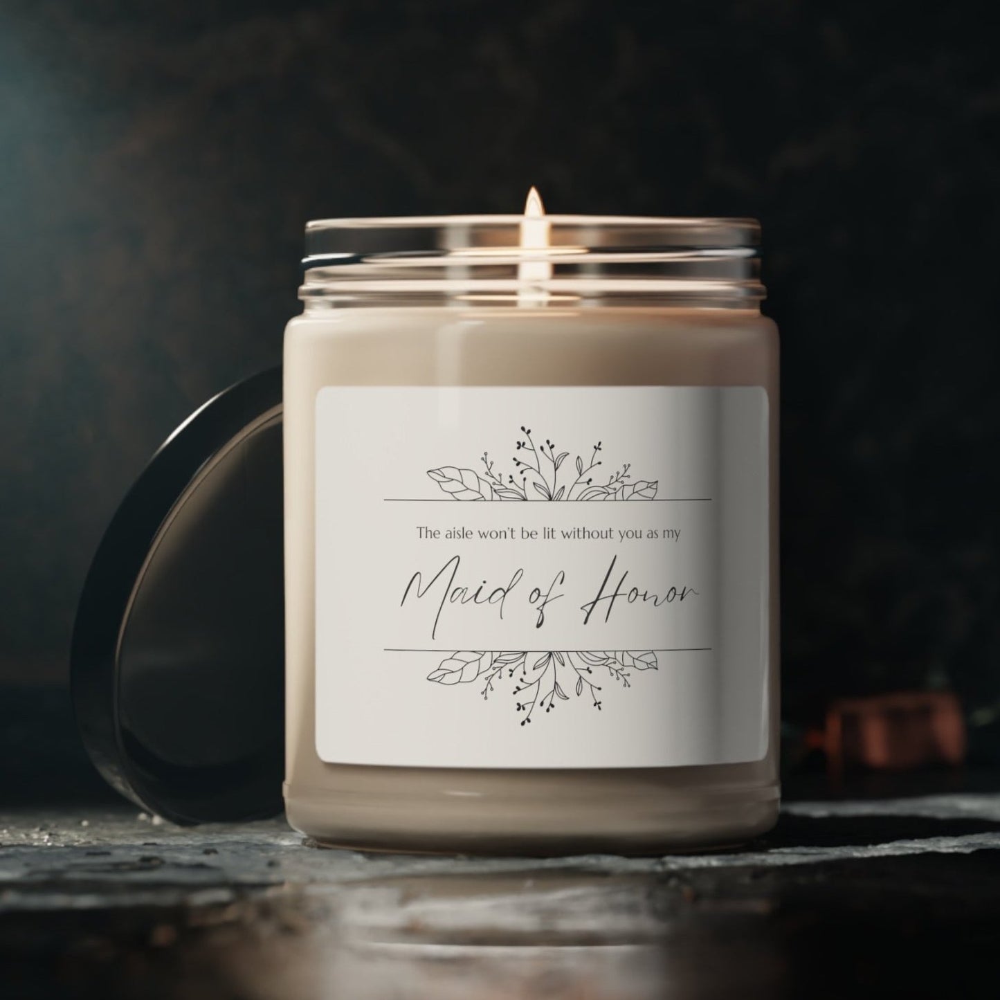 A picture of our 9 Oz Soy Candle with the label that says The Aisle wont be lit without you as my Maid of Honor. There are Beautiful floral border drawing on the top and bottom of the saying, in simple and elegant black ink color.