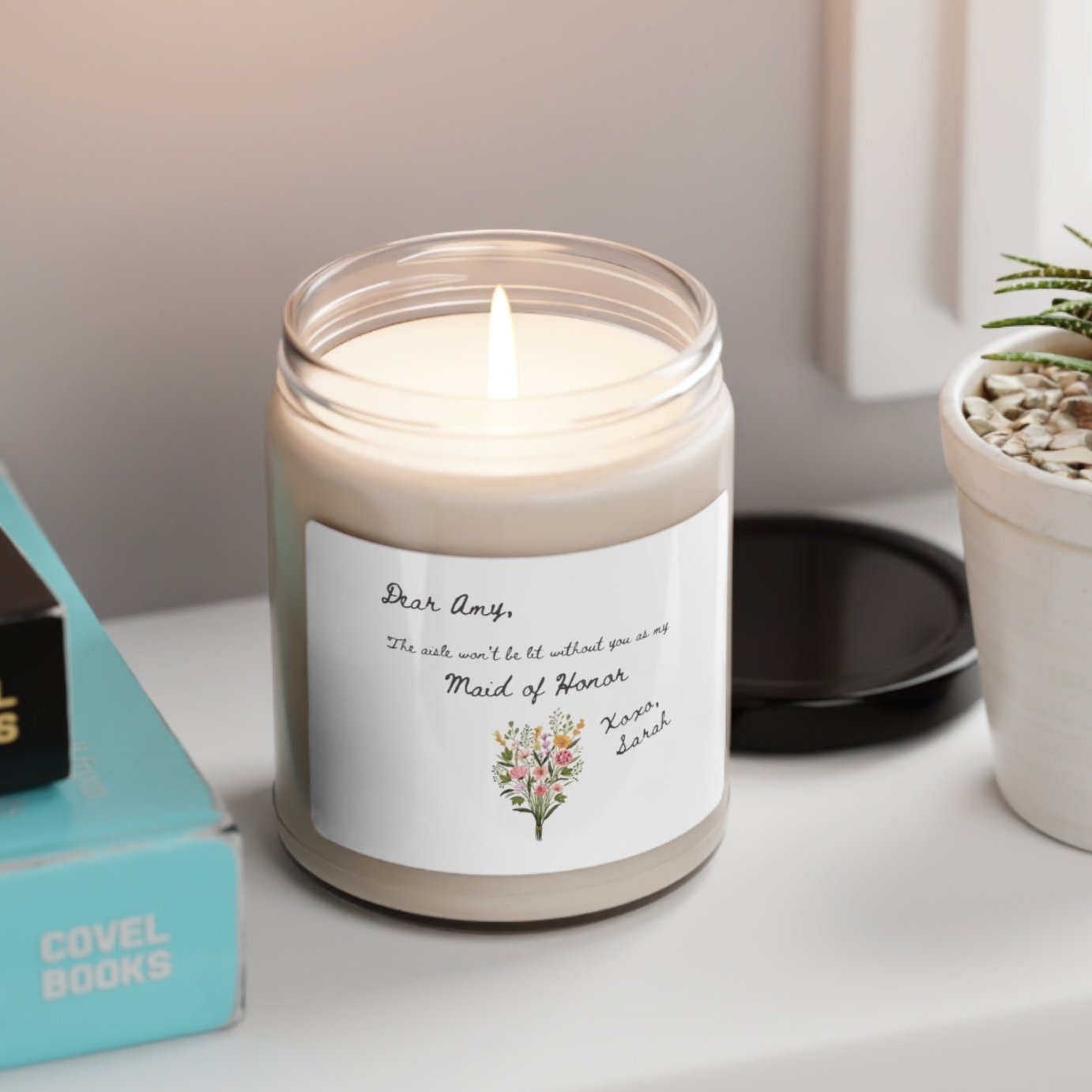 A picture of our 9 Oz Soy Candle with the label that says Dear Amy, The Aisle wont be lit without you as my Maid of Honor. Xoxo, Sarah. The names can be personalized. It is also adorned with an image of a beautiful bouquet of flowers.