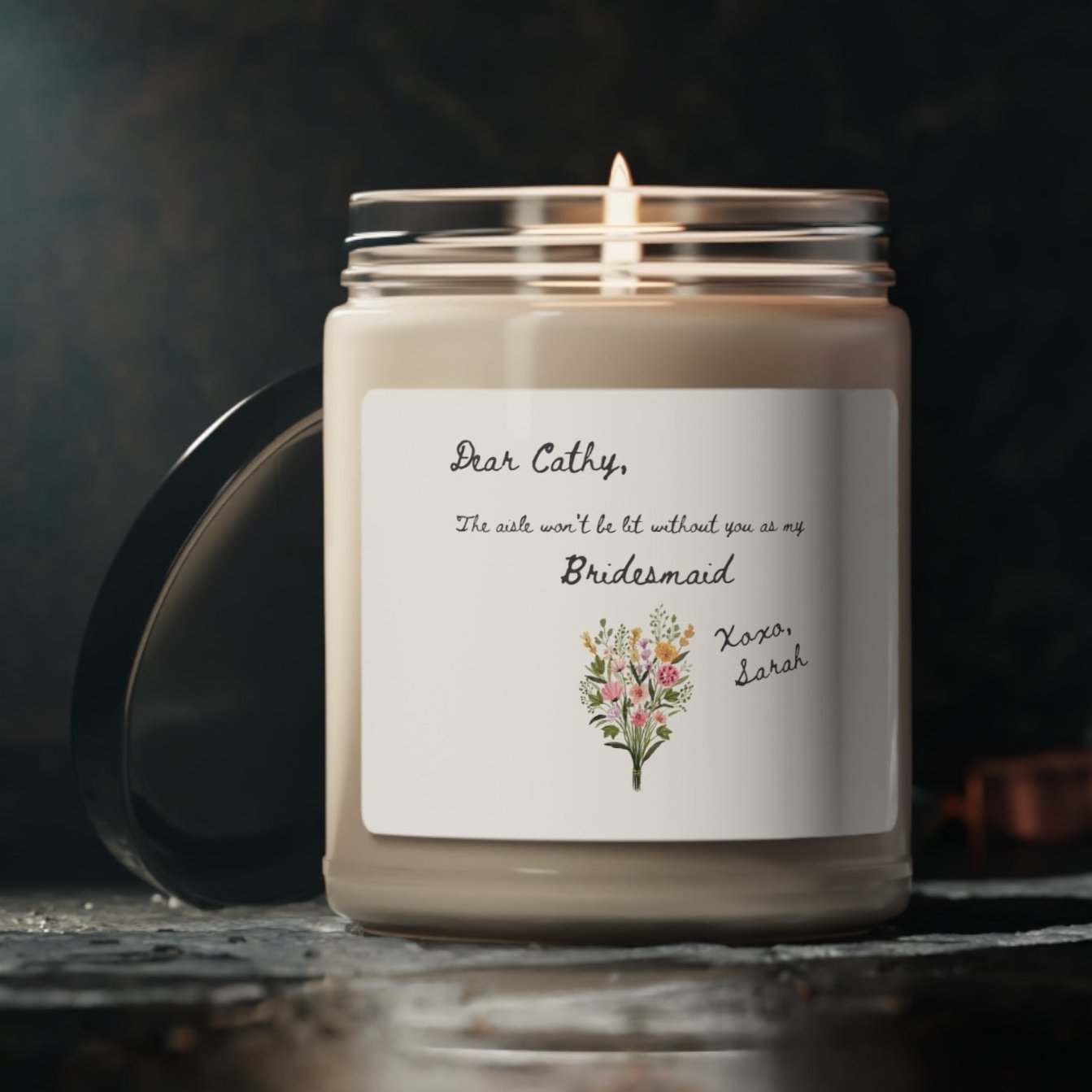 A picture of our 9 Oz Soy Candle with the label that says Dear Cathy, The Aisle wont be lit without you as my Bridesmaid. Xoxo, Sarah. The names can be personalized. It is also adorned with an image of a beautiful bouquet of flowers.