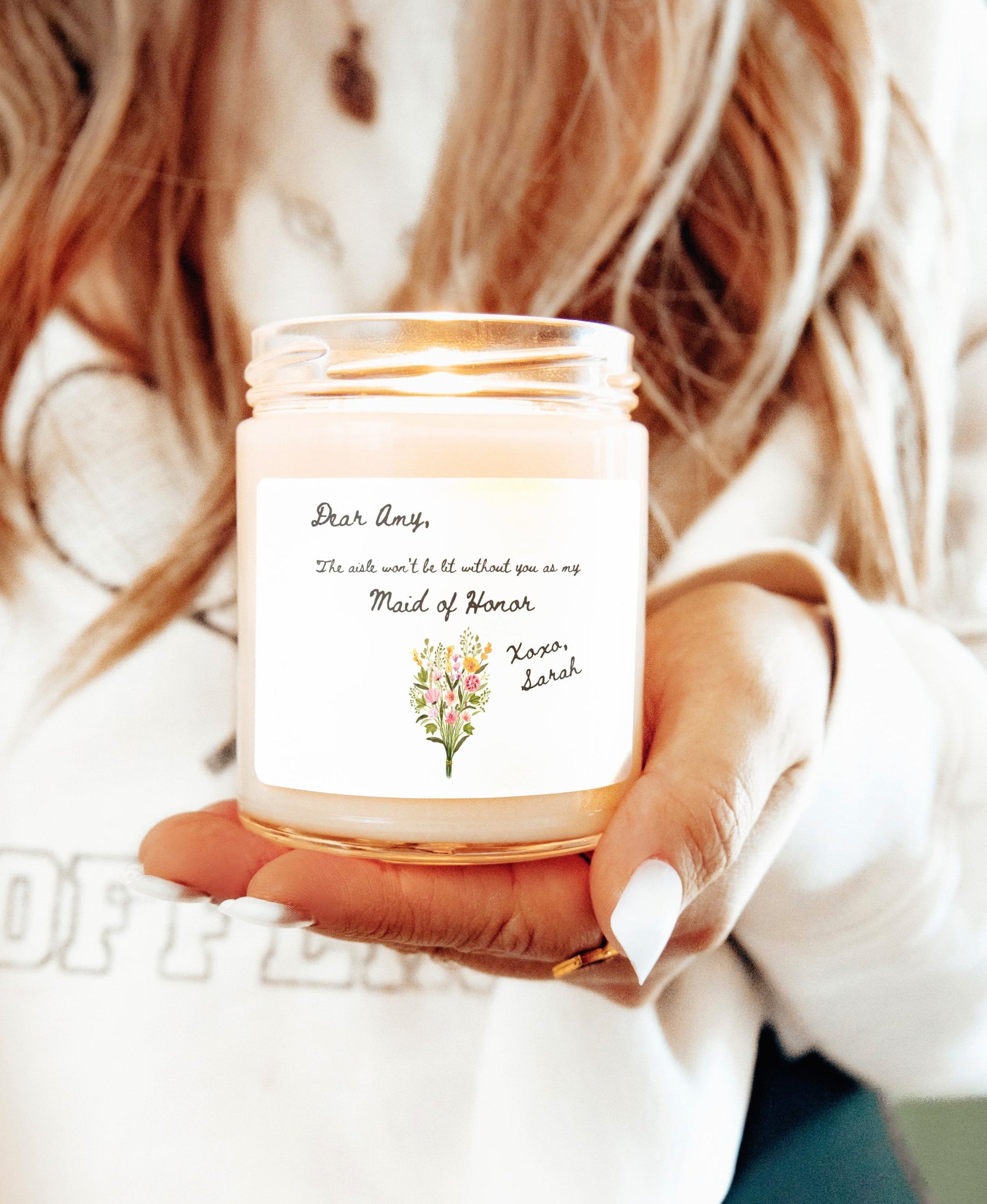 A picture of our 9 Oz Soy Candle with the label that says Dear Amy, The Aisle wont be lit without you as my Maid of Honor. Xoxo, Sarah. The names can be personalized. It is also adorned with an image of a beautiful bouquet of flowers.