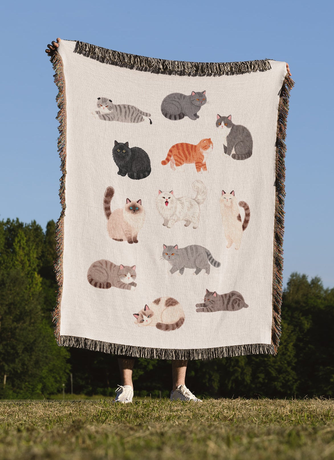 A person standing outdoors in a grassy field under a clear blue sky holds up a large cream-colored throw blanket featuring multiple illustrated cats in various colors. The blanket has dark fringe on all sides.