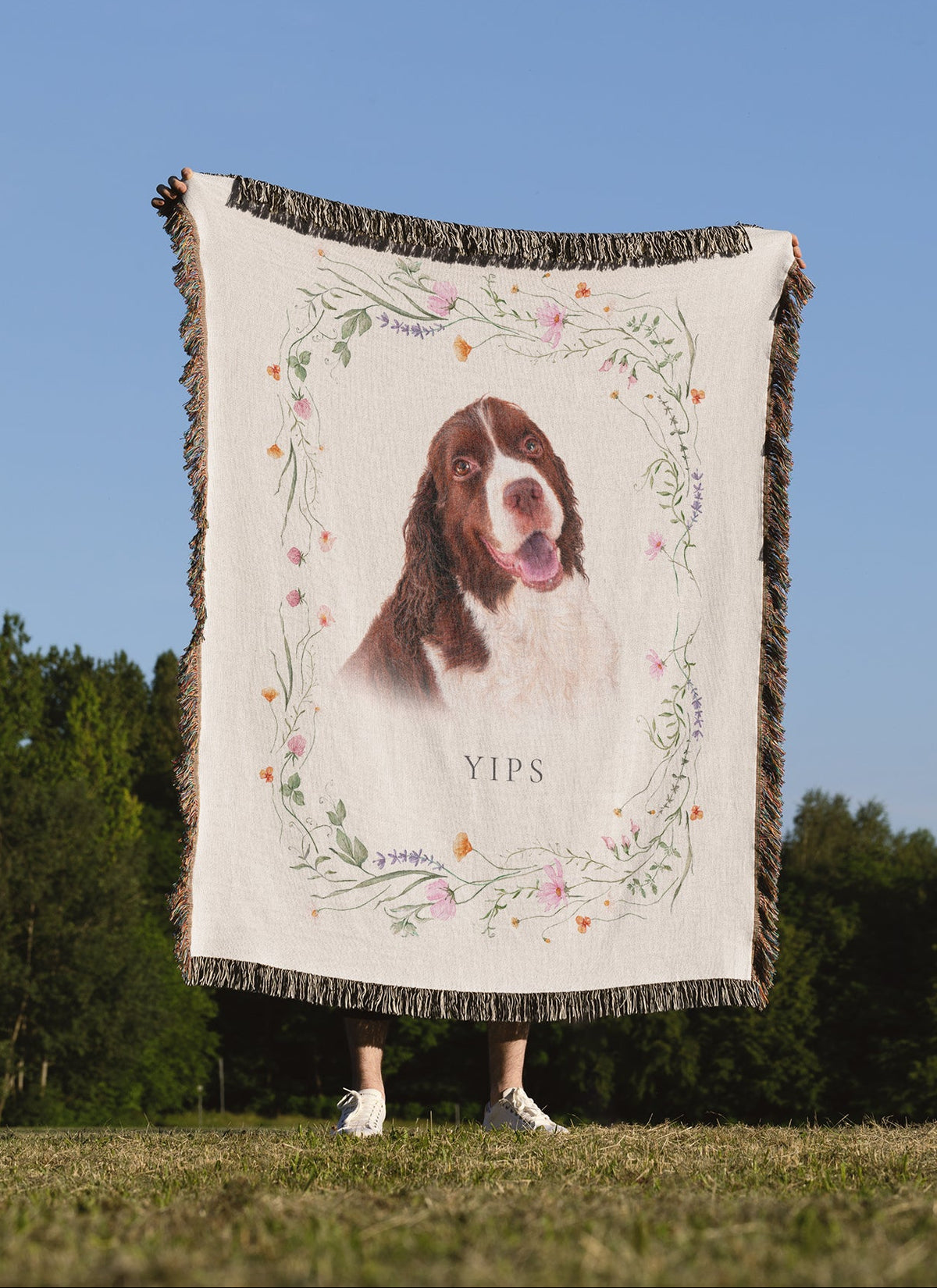 A person standing outdoors in a grassy field under a blue sky holds up a large woven throw blanket featuring a printed portrait of a dog framed by a dainty floral border.