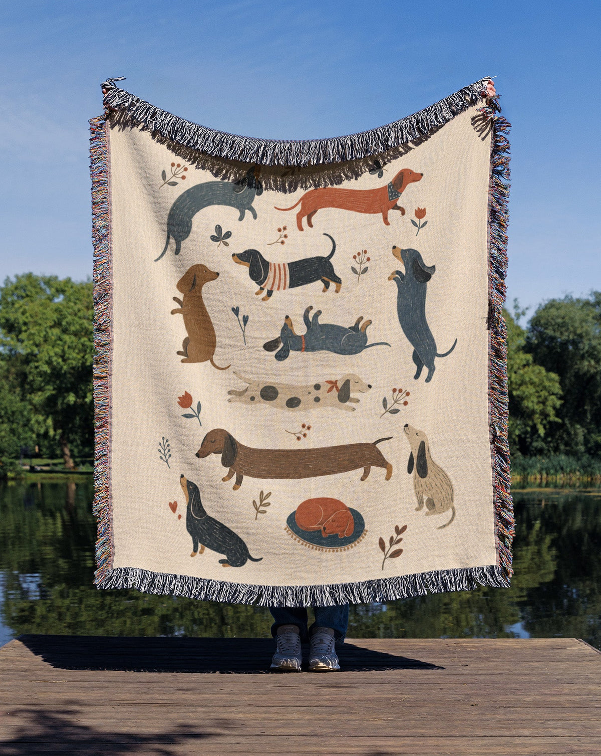 A large woven throw blanket with fringe, featuring a playful pattern of dachshund dogs in various poses, held up by a person standing on a wooden dock with a lake and trees in the background.