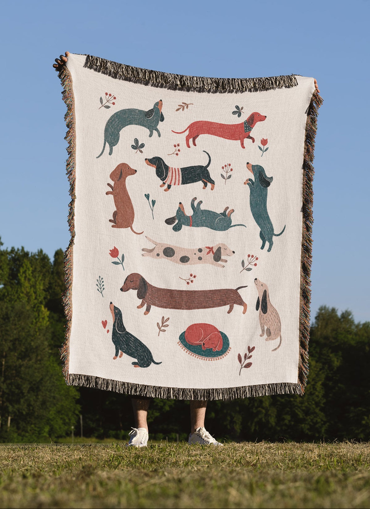 A person outdoors holding up a large woven throw blanket featuring illustrated dachshunds in multiple colors on an off-white background, with trees and blue sky behind them.