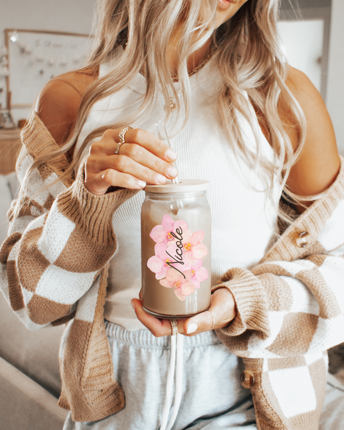 A picture of a smiling woman holding our 16 Oz Sipper Glass filled with Iced Coffee, with the name Nicole and a pretty Pink Orchid Flower background. Name is customizable and there are 4 Flower background options to choose from.