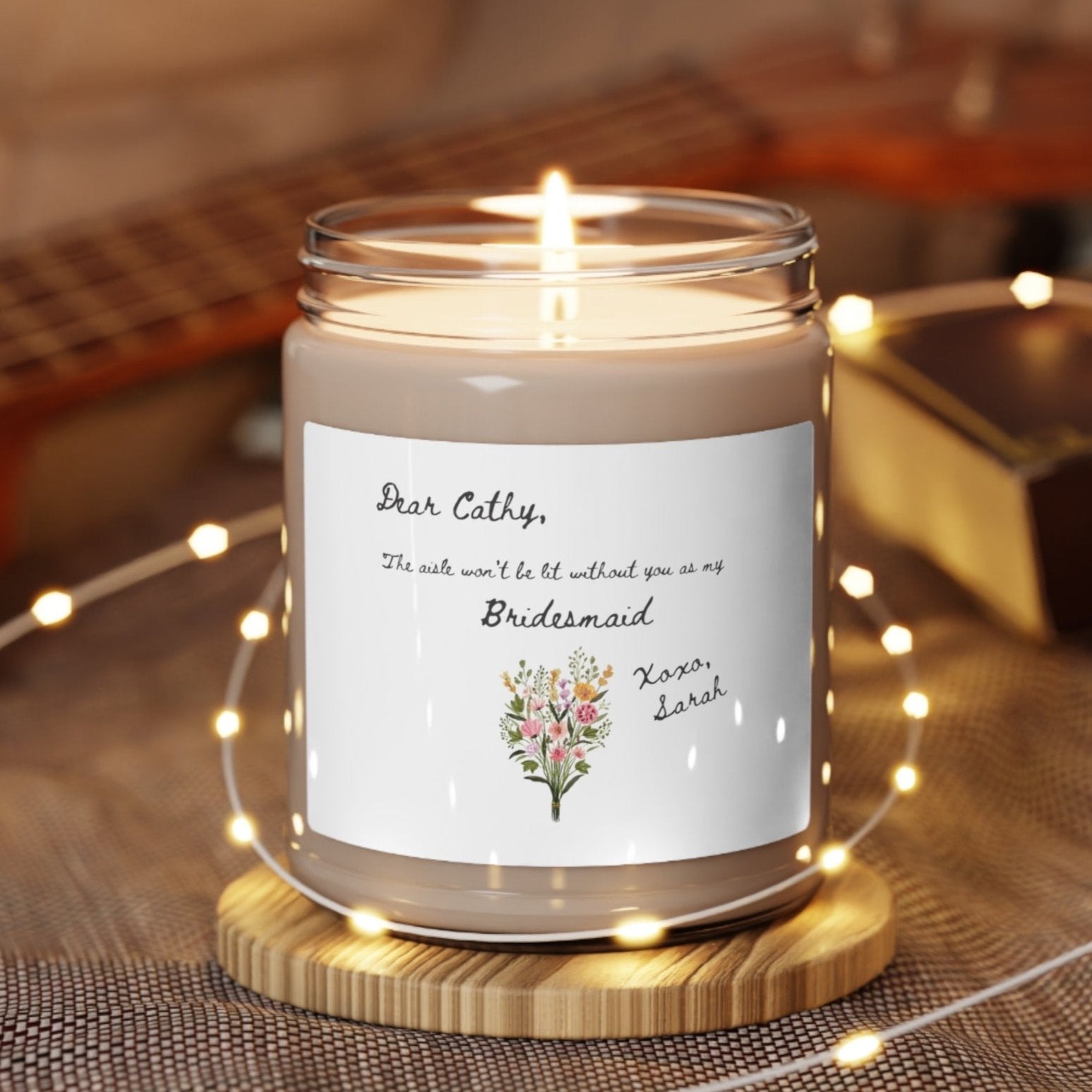 A picture of our 9 Oz Soy Candle with the label that says Dear Cathy, The Aisle wont be lit without you as my Bridesmaid. Xoxo, Sarah. The names can be personalized. It is also adorned with an image of a beautiful bouquet of flowers.