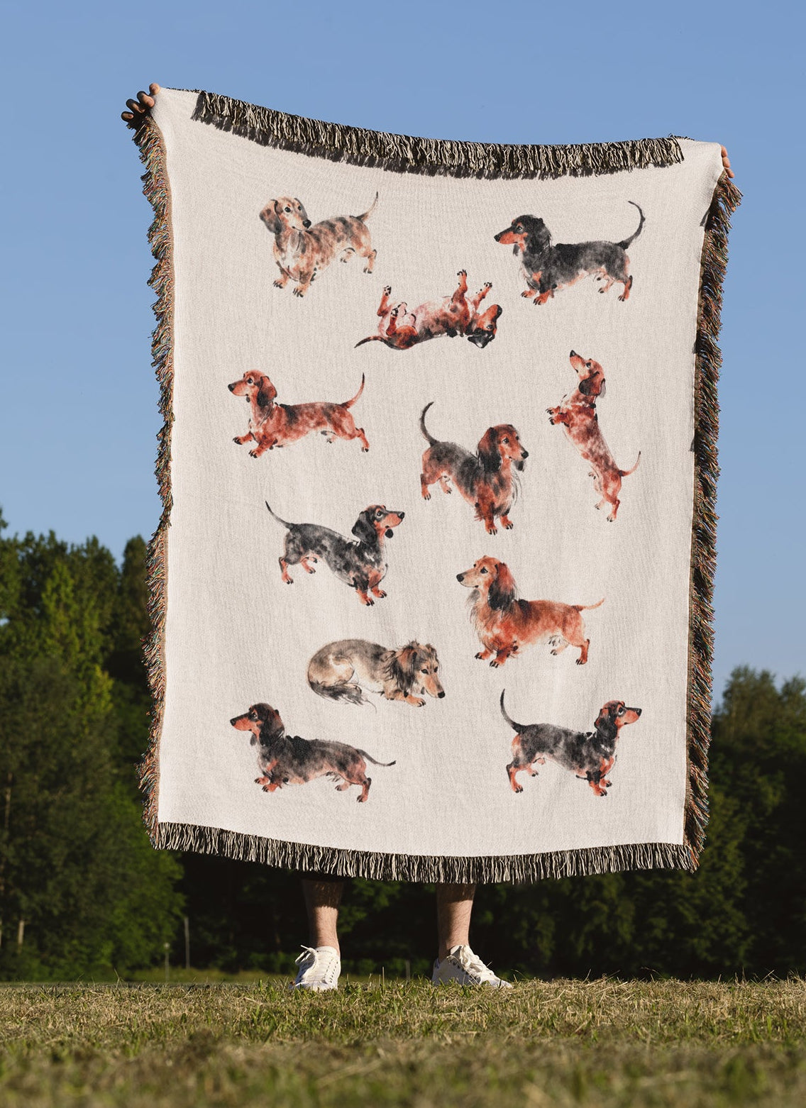 A person outdoors holding up a large cream-colored woven blanket with fringed edges and a print of multiple dachshund dogs in various poses, against a blue sky and green trees.